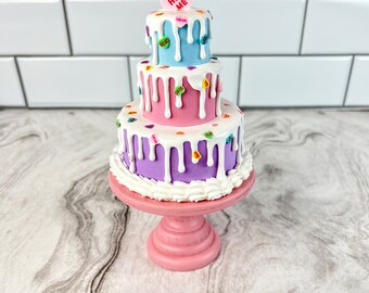 May include: A three-tiered cake decorated with colourful candy hearts and white frosting. The top tier is light blue, the middle tier is pink, and the bottom tier is purple. The cake is on a pink cake stand. The top of the cake has a candy heart that says "Hug Me".