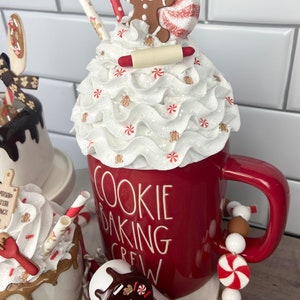 May include: A red mug with white lettering that reads "Cookie Baking Crew" topped with a white whipped cream and a gingerbread man, candy cane, and rolling pin. The mug is on a tiered stand with white and brown beads and a smaller white mug with brown chocolate sauce and sprinkles.
