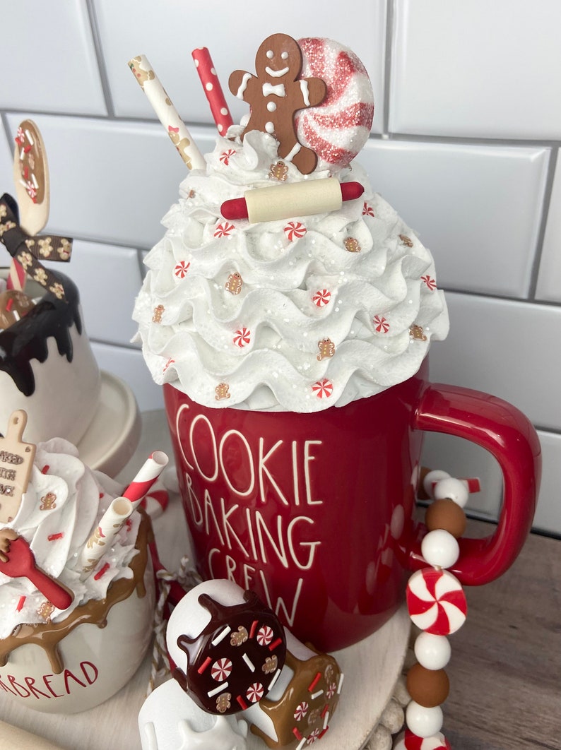 May include: A red mug with white lettering that reads "Cookie Baking Crew" topped with a white whipped cream and a gingerbread man, candy cane, and rolling pin. The mug is on a tiered stand with white and brown beads and a smaller white mug with brown chocolate sauce and sprinkles.