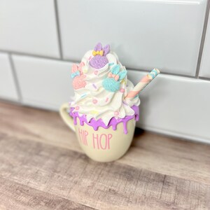 K&ouml;nnte beinhalten: Eine wei&szlig;e Keramiktasse mit einem violetten Tropf-Glanz und dem Text "HIP HOP" in Pink. Die Tasse ist mit Schlagsahne, Streuseln und drei pastellfarbenen Hasenfiguren verziert.