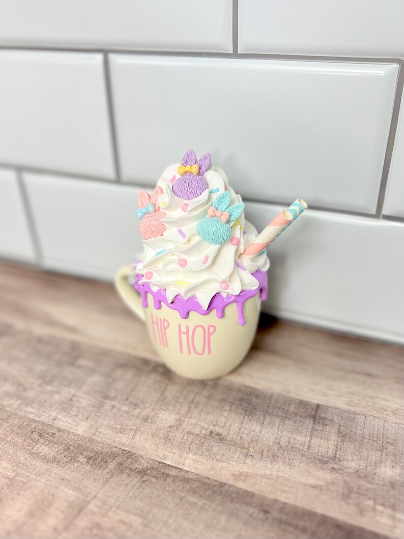 K&ouml;nnte beinhalten: Eine wei&szlig;e Keramiktasse mit einem violetten Tropf-Glanz und dem Text "HIP HOP" in Pink. Die Tasse ist mit Schlagsahne, Streuseln und drei pastellfarbenen Hasenfiguren verziert.