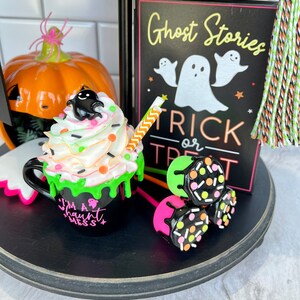 May include: A Halloween-themed tiered tray with a black and white sign that reads "Ghost Stories Trick or Treat", a green and black mug with white frosting and sprinkles that says "I'm a Haunt Mess", and three pink and black lollipop-style treats with sprinkles.