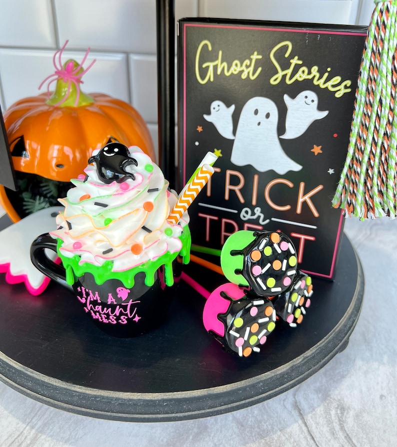 May include: A Halloween-themed tiered tray with a black and white sign that reads "Ghost Stories Trick or Treat", a green and black mug with white frosting and sprinkles that says "I'm a Haunt Mess", and three pink and black lollipop-style treats with sprinkles.