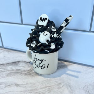 May include: A black and white Halloween themed mug with a black and white whipped cream topping, a black and white striped straw, and a ghost and tombstone decoration. The mug says "hey boo!"