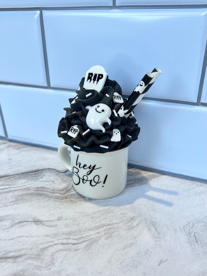 May include: A black and white Halloween themed mug with a black and white whipped cream topping, a black and white striped straw, and a ghost and tombstone decoration. The mug says "hey boo!"
