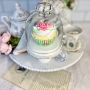 May include: A white cupcake with pink and white frosting and a green cupcake liner is displayed under a glass dome on a white tiered stand. The cupcake is decorated with a pink rose.