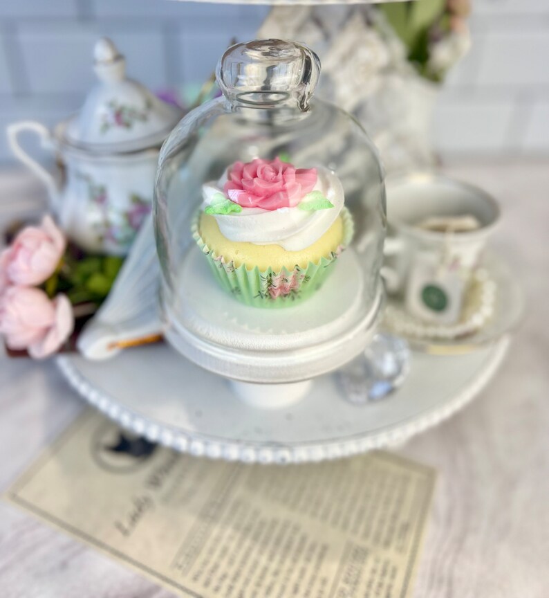 May include: A white cupcake with pink and white frosting and a green cupcake liner is displayed under a glass dome on a white tiered stand. The cupcake is decorated with a pink rose.