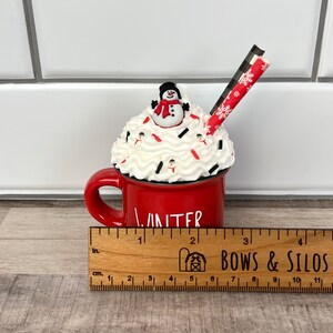 Op de afbeelding: Rode keramische mok met witte slagroom en een sneeuwpop erop. De mok heeft de tekst "Winter" en een rood-wit gestreepte rietje. De mok staat op een houten liniaal met de tekst "Bows & Silos".