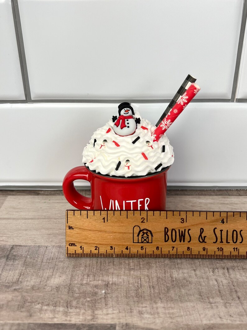 Op de afbeelding: Rode keramische mok met witte slagroom en een sneeuwpop erop. De mok heeft de tekst "Winter" en een rood-wit gestreepte rietje. De mok staat op een houten liniaal met de tekst "Bows & Silos".