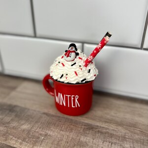 Op de afbeelding: Rode keramische mok met het woord "WINTER" aan de zijkant, gevuld met witte slagroom, rode en witte sprinkles en een sneeuwpopfiguur bovenop. Een rood-wit gestreepte rietje steekt uit de slagroom.