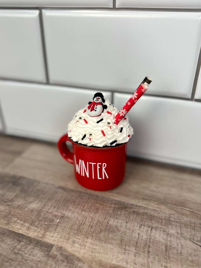 Op de afbeelding: Rode keramische mok met het woord "WINTER" aan de zijkant, gevuld met witte slagroom, rode en witte sprinkles en een sneeuwpopfiguur bovenop. Een rood-wit gestreepte rietje steekt uit de slagroom.