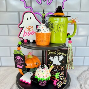 May include: A two-tiered black tiered tray decorated for Halloween. The top tier features a green mug with the word "Toxic" on it, a cupcake with orange and black frosting, a ghost with the words "Hey Boo" on it, and a purple neon bat. The bottom tier features a pumpkin, a ghost, a green mug with white frosting and a black spider on top, a sign that says "Ghost Stories Trick or Treat", and a green and black tassel.