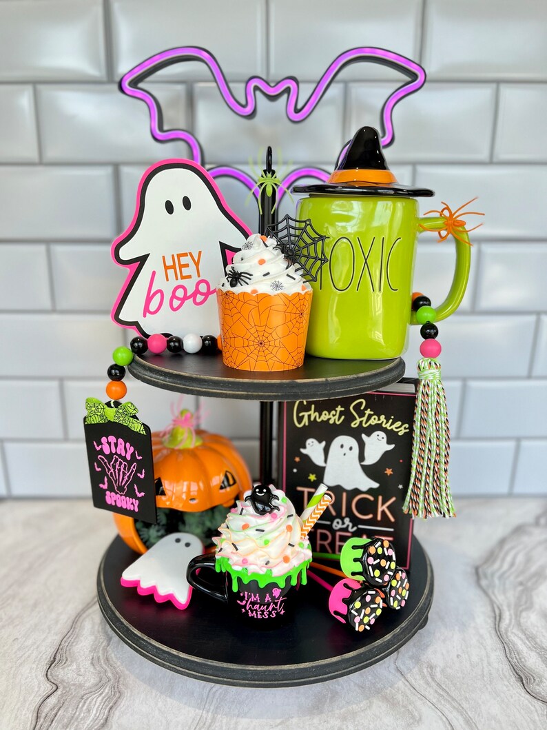 May include: A two-tiered black tiered tray decorated for Halloween. The top tier features a green mug with the word "Toxic" on it, a cupcake with orange and black frosting, a ghost with the words "Hey Boo" on it, and a purple neon bat. The bottom tier features a pumpkin, a ghost, a green mug with white frosting and a black spider on top, a sign that says "Ghost Stories Trick or Treat", and a green and black tassel.