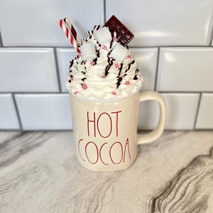 May include: White ceramic mug with red lettering that says "HOT COCOA". The mug is topped with whipped cream, chocolate pieces, marshmallows, and red and white candy canes.