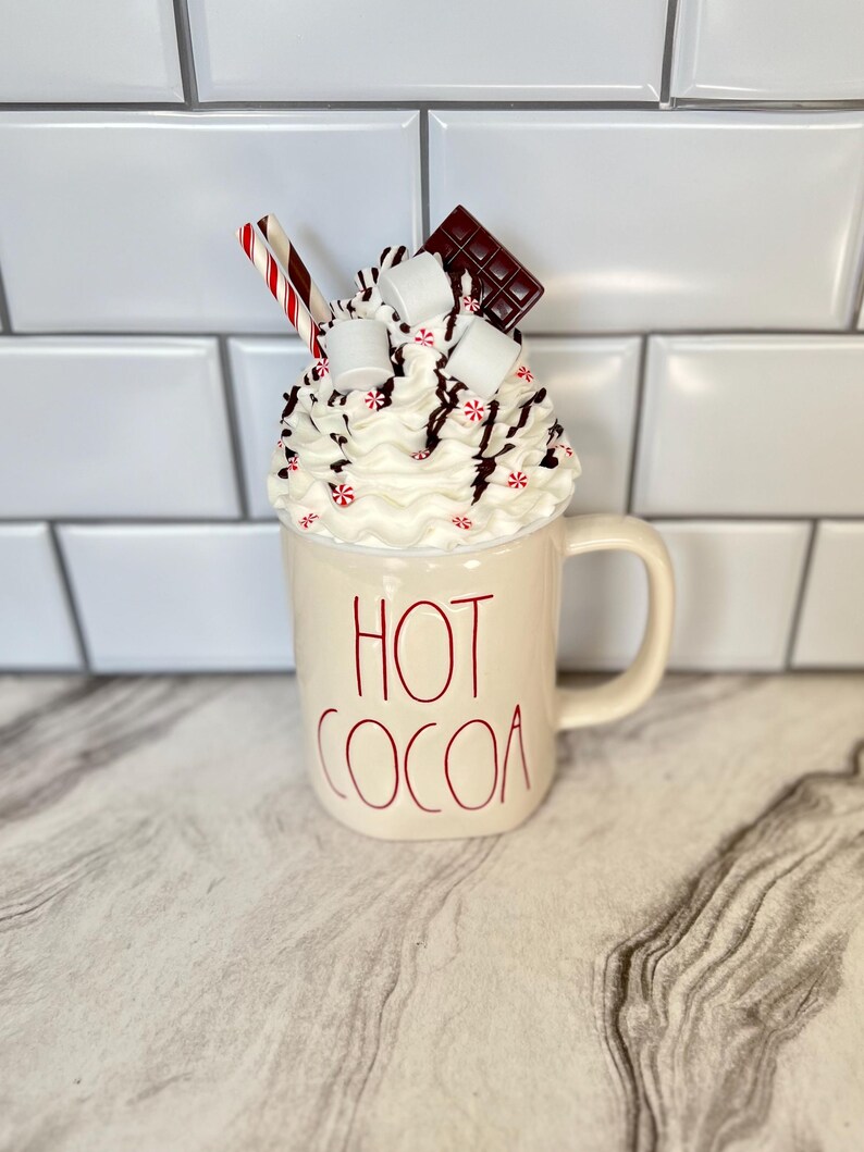 May include: White ceramic mug with red lettering that says "HOT COCOA". The mug is topped with whipped cream, chocolate pieces, marshmallows, and red and white candy canes.