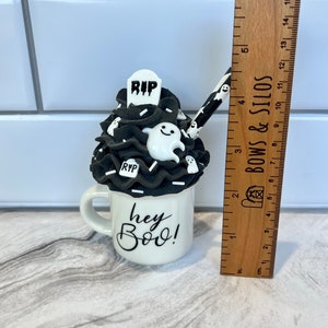 May include: A white ceramic mug with a black and white Halloween-themed design. The mug has a black and white frosting-like topping with a ghost, a tombstone that says "RIP", and white sprinkles. The mug says "hey Boo!" in black lettering.