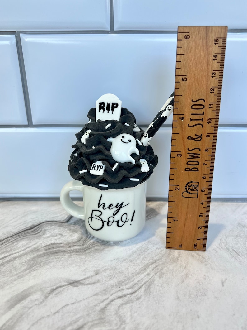 May include: A white ceramic mug with a black and white Halloween-themed design. The mug has a black and white frosting-like topping with a ghost, a tombstone that says "RIP", and white sprinkles. The mug says "hey Boo!" in black lettering.