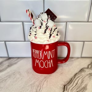 May include: Red ceramic mug with white lettering that reads "Peppermint Mocha". The mug is topped with whipped cream, chocolate shavings, marshmallows, and a red and white striped straw.
