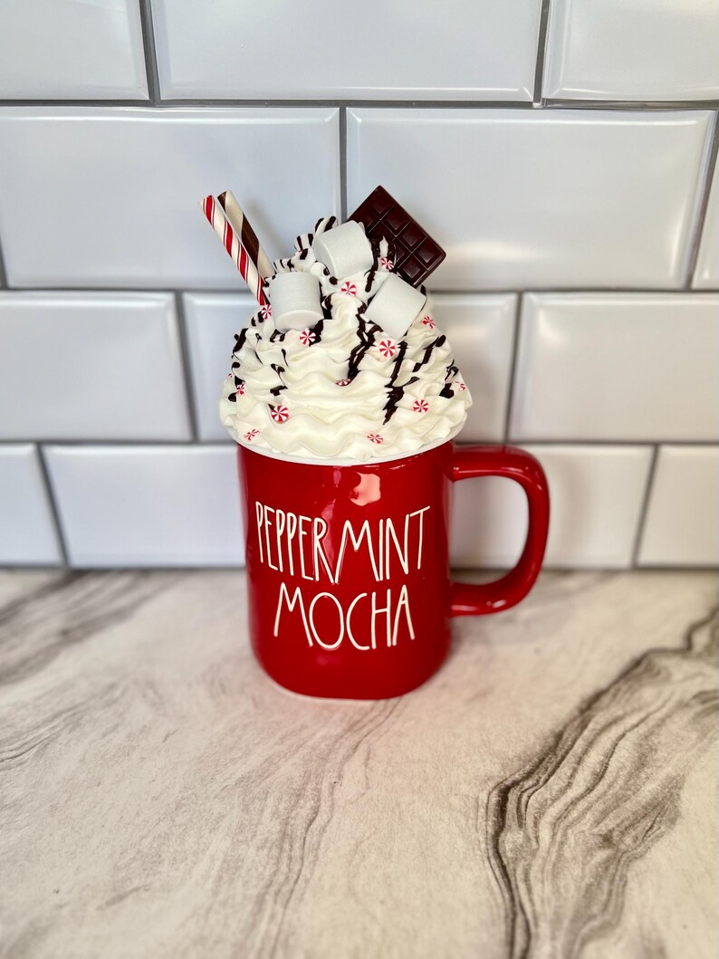 May include: Red ceramic mug with white lettering that reads "Peppermint Mocha". The mug is topped with whipped cream, chocolate shavings, marshmallows, and a red and white striped straw.