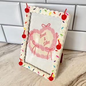 May include: White picture frame with a frosting and sprinkles design. The frame has red cherries around the edge. The frame has a pink heart with the text "Bows & Silos" inside.