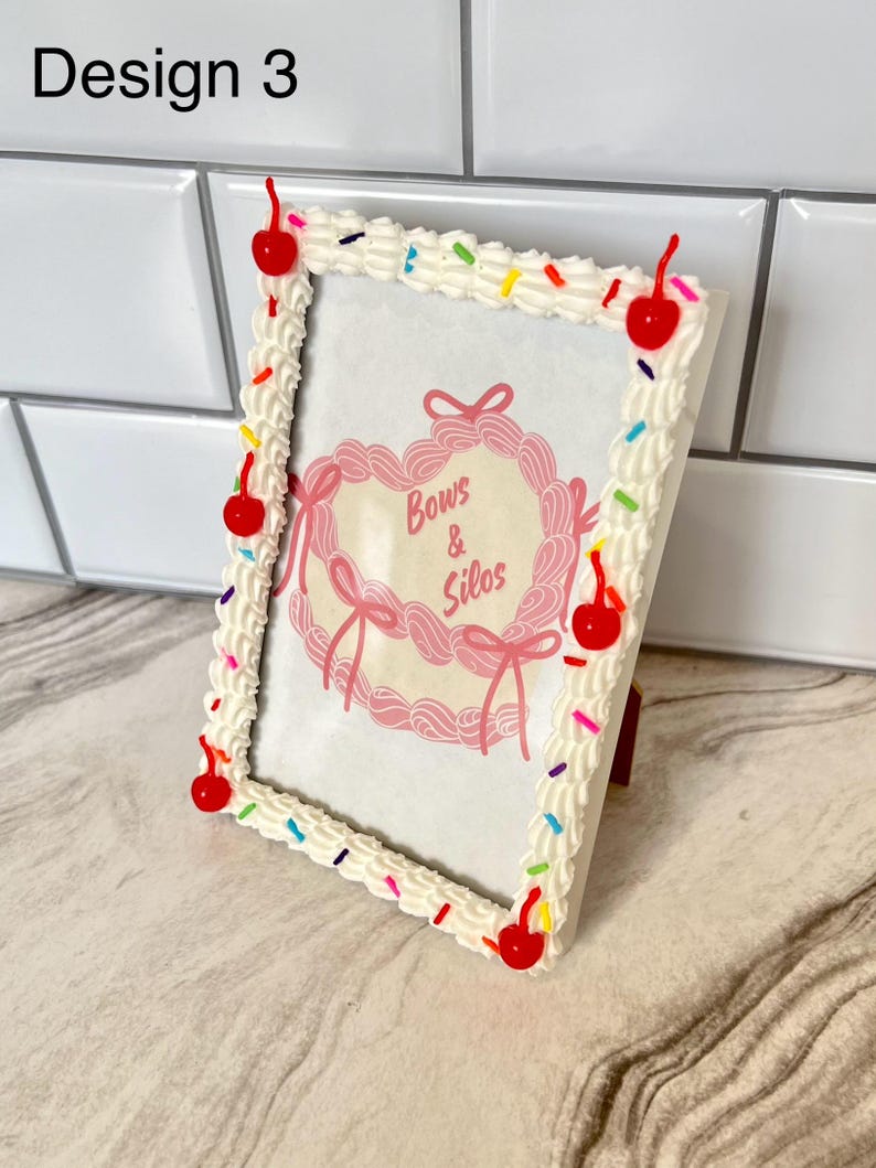 May include: White picture frame with a frosting and sprinkles design. The frame has red cherries around the edge. The frame has a pink heart with the text "Bows & Silos" inside.
