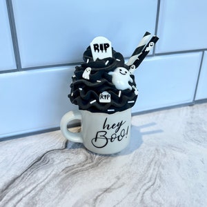 May include: A white ceramic mug with black lettering that says "hey boo!"  The mug is filled with a black and white whipped cream topping with a tombstone, ghosts, and sprinkles.  Two black and white striped straws are sticking out of the whipped cream.