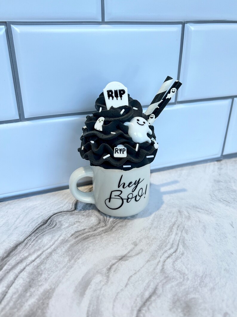 May include: A white ceramic mug with black lettering that says "hey boo!"  The mug is filled with a black and white whipped cream topping with a tombstone, ghosts, and sprinkles.  Two black and white striped straws are sticking out of the whipped cream.