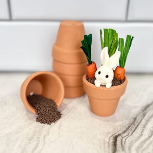 May include: A miniature terracotta flower pot with a white bunny figurine, orange carrots, and green leaves. Another pot is tipped over, spilling brown soil. Several other pots are stacked in the background. The scene is set on a white and gray surface.