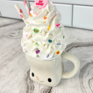 May include: A white ceramic mug with a smiling face, filled with whipped cream and topped with colorful candy hearts, sprinkles, and two pink and white striped straws. The candy hearts have messages like "Be Mine", "Love You", and "XOXO".