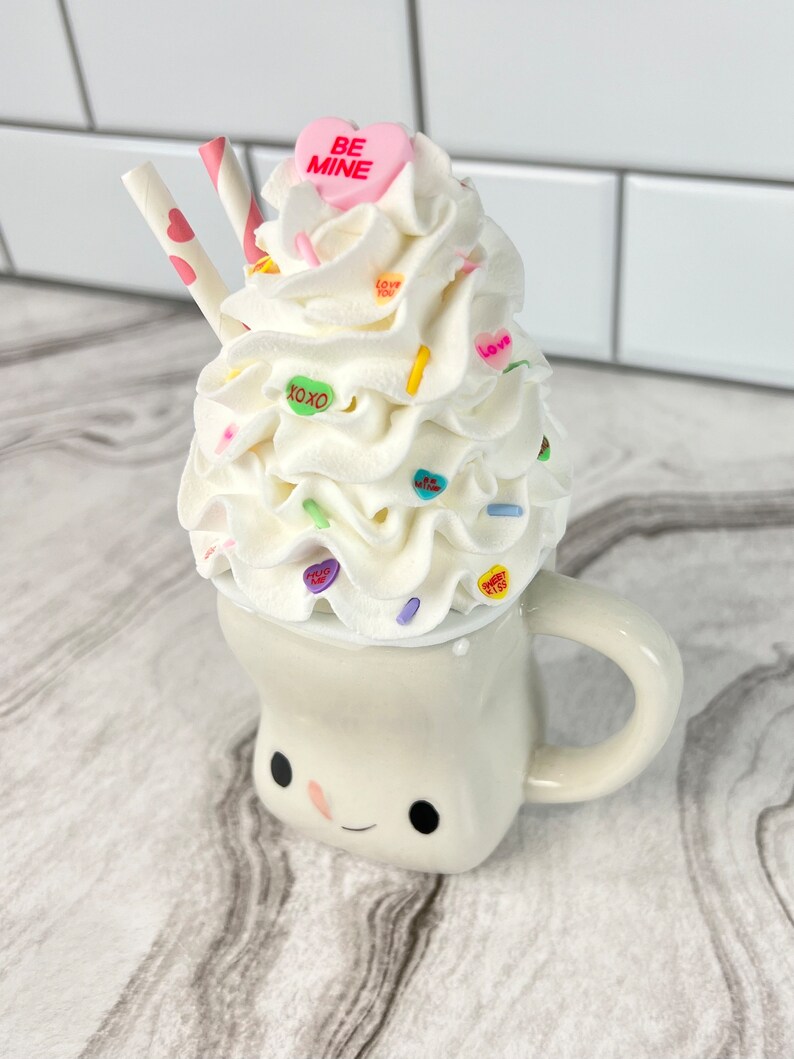 May include: A white ceramic mug with a smiling face, filled with whipped cream and topped with colorful candy hearts, sprinkles, and two pink and white striped straws. The candy hearts have messages like "Be Mine", "Love You", and "XOXO".