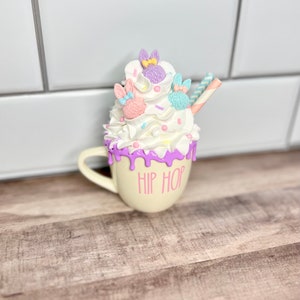 K&ouml;nnte beinhalten: Eine wei&szlig;e Keramiktasse mit einem violetten Tropfendesign und dem Text "HIP HOP" an der Seite. Die Tasse ist mit Schlagsahne gef&uuml;llt und mit pastellfarbenen, kaninchenf&ouml;rmigen S&uuml;&szlig;igkeiten und einem gestreiften Strohhalm verziert.