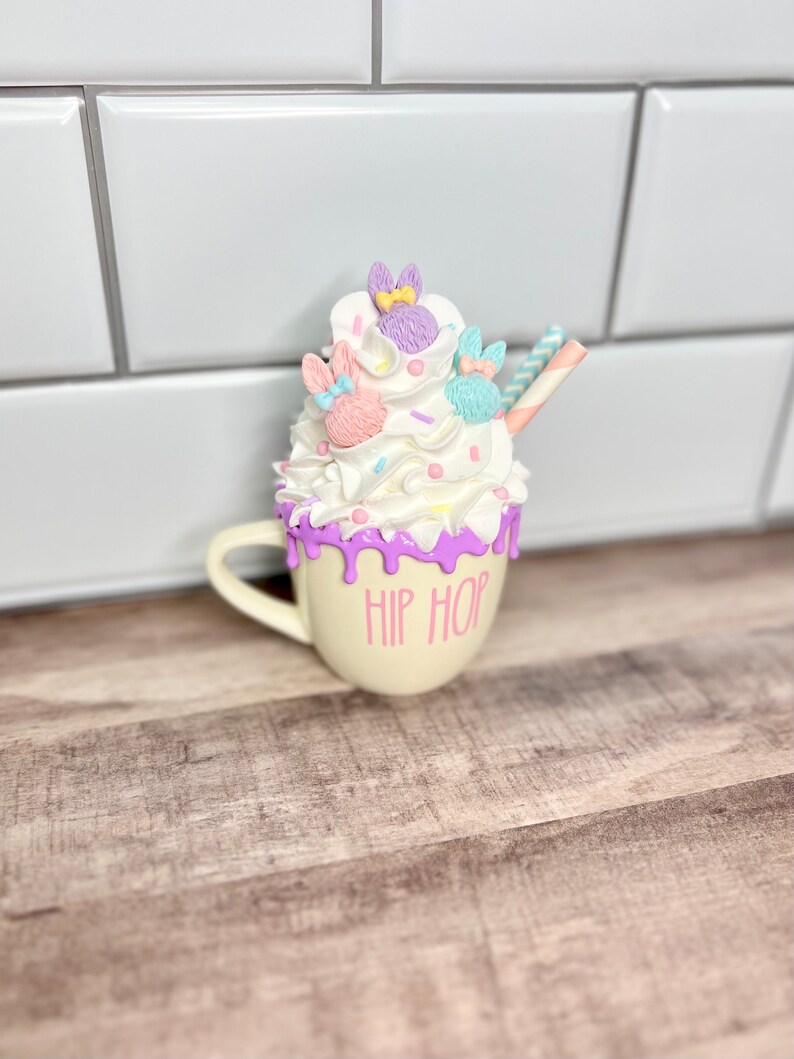 K&ouml;nnte beinhalten: Eine wei&szlig;e Keramiktasse mit einem violetten Tropfendesign und dem Text "HIP HOP" an der Seite. Die Tasse ist mit Schlagsahne gef&uuml;llt und mit pastellfarbenen, kaninchenf&ouml;rmigen S&uuml;&szlig;igkeiten und einem gestreiften Strohhalm verziert.
