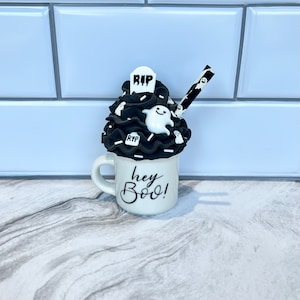 May include: White ceramic mug with black lettering that says "hey boo!"  The mug is filled with black and white frosting and topped with a white ghost and a black tombstone that says "RIP". A black and white striped straw is sticking out of the frosting.