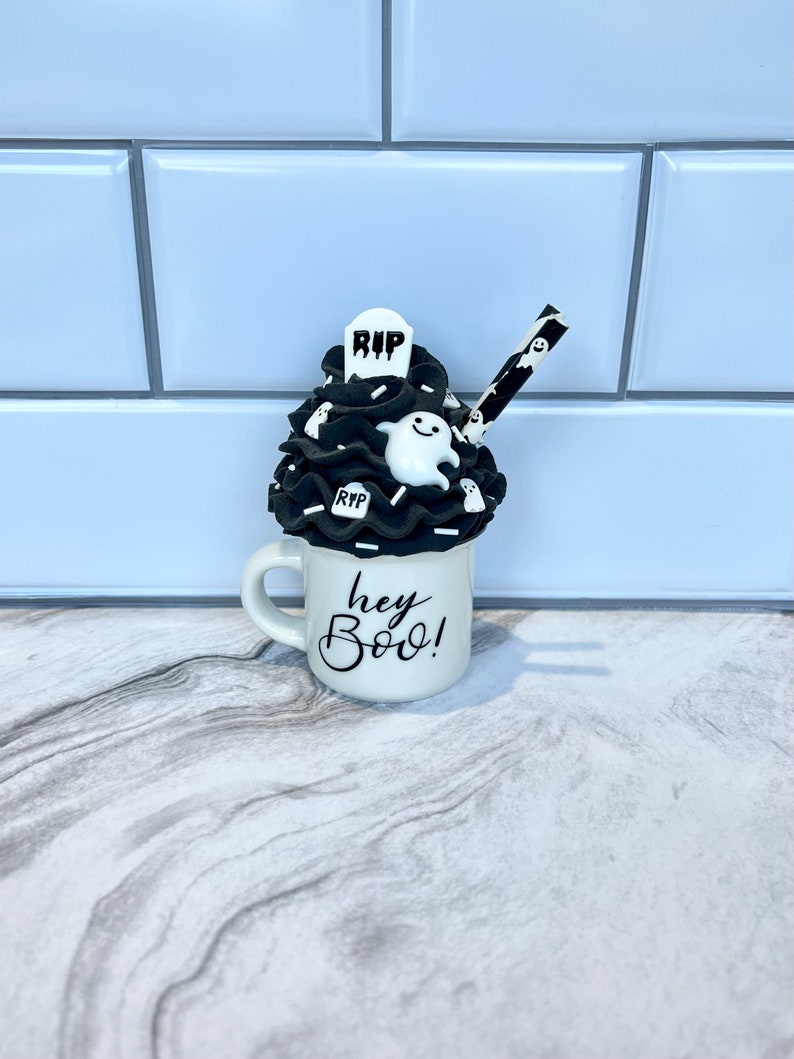 May include: White ceramic mug with black lettering that says "hey boo!"  The mug is filled with black and white frosting and topped with a white ghost and a black tombstone that says "RIP". A black and white striped straw is sticking out of the frosting.