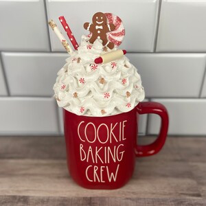 May include: Red ceramic mug with a white whipped cream and candy cane topping. A gingerbread man and a rolling pin are on top of the whipped cream. The mug says "Cookie Baking Crew".