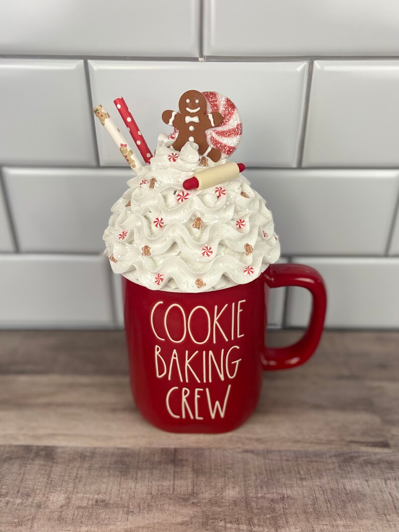 May include: Red ceramic mug with a white whipped cream and candy cane topping. A gingerbread man and a rolling pin are on top of the whipped cream. The mug says "Cookie Baking Crew".