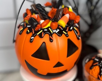 May include: Orange pumpkin-shaped candy bucket with a black jack-o'-lantern face. The bucket is filled with assorted Halloween candy, including candy corn, chocolate, and licorice. The bucket has a black wire handle.