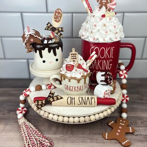 May include: A white tiered tray decorated with a gingerbread house, a red mug with white frosting and a gingerbread man, a white mug with chocolate sauce and a gingerbread man, a red rolling pin with the text "Oh Snap!" and a red and white beaded garland.