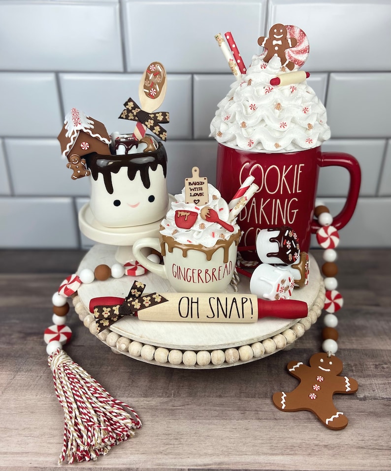 May include: A white tiered tray decorated with a gingerbread house, a red mug with white frosting and a gingerbread man, a white mug with chocolate sauce and a gingerbread man, a red rolling pin with the text "Oh Snap!" and a red and white beaded garland.