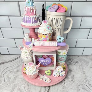 May include: A pink tiered tray decorated with Valentine's Day themed items, including a mini cake, a cupcake, a mug, a hot chocolate mug, and a picture frame. The tray is decorated with candy hearts, sprinkles, and other Valentine's Day decorations. The text on the mug reads "Candy Hearts".