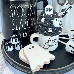 May include: A black mug with the text "Spooky Season" on it, a white mug with the text "Hey Boo!" on it, a black and white ghost-themed cupcake, and a black tray with white ghost-shaped cookies.