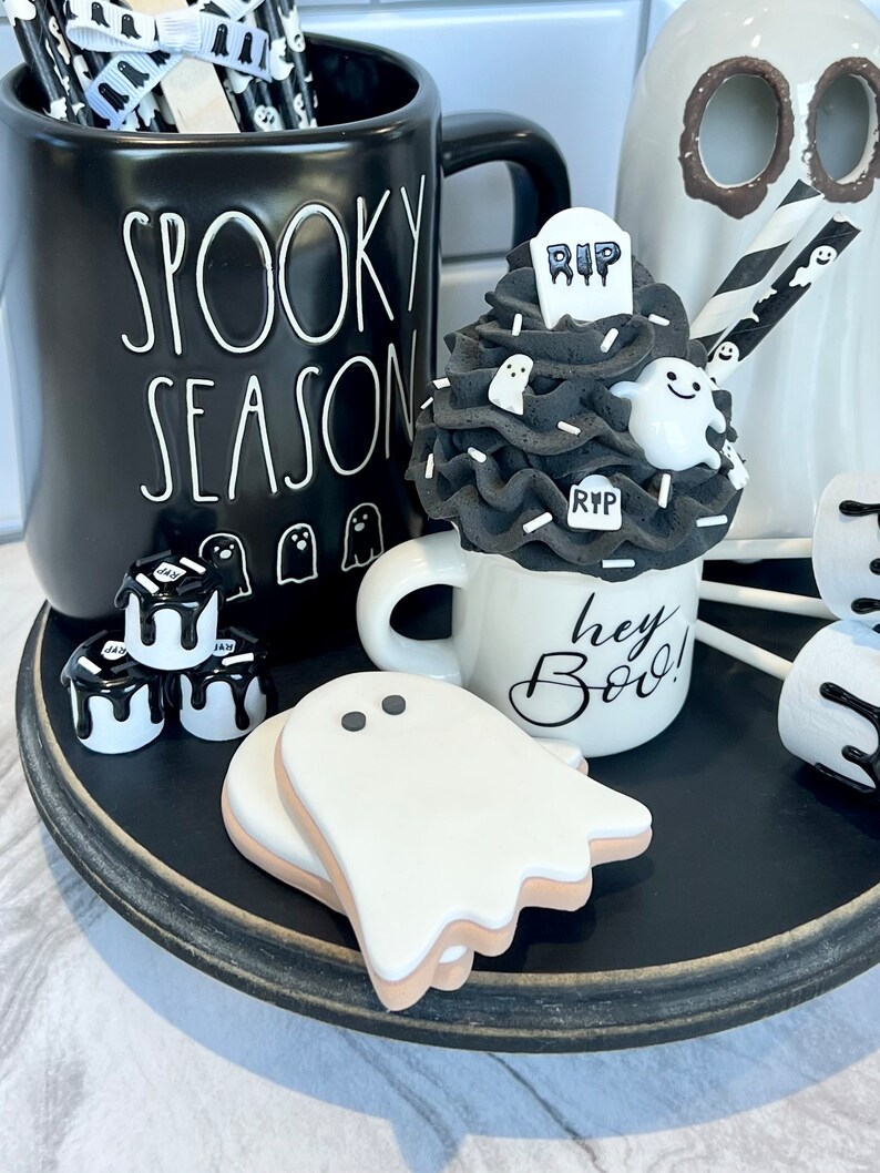 May include: A black mug with the text "Spooky Season" on it, a white mug with the text "Hey Boo!" on it, a black and white ghost-themed cupcake, and a black tray with white ghost-shaped cookies.