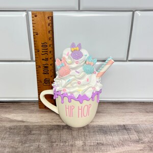 K&ouml;nnte beinhalten: Eine wei&szlig;e Keramiktasse mit rosa und lila Frosting-Design und einem gestreiften Strohhalm. Auf der Tasse steht "HIP HOP" geschrieben.