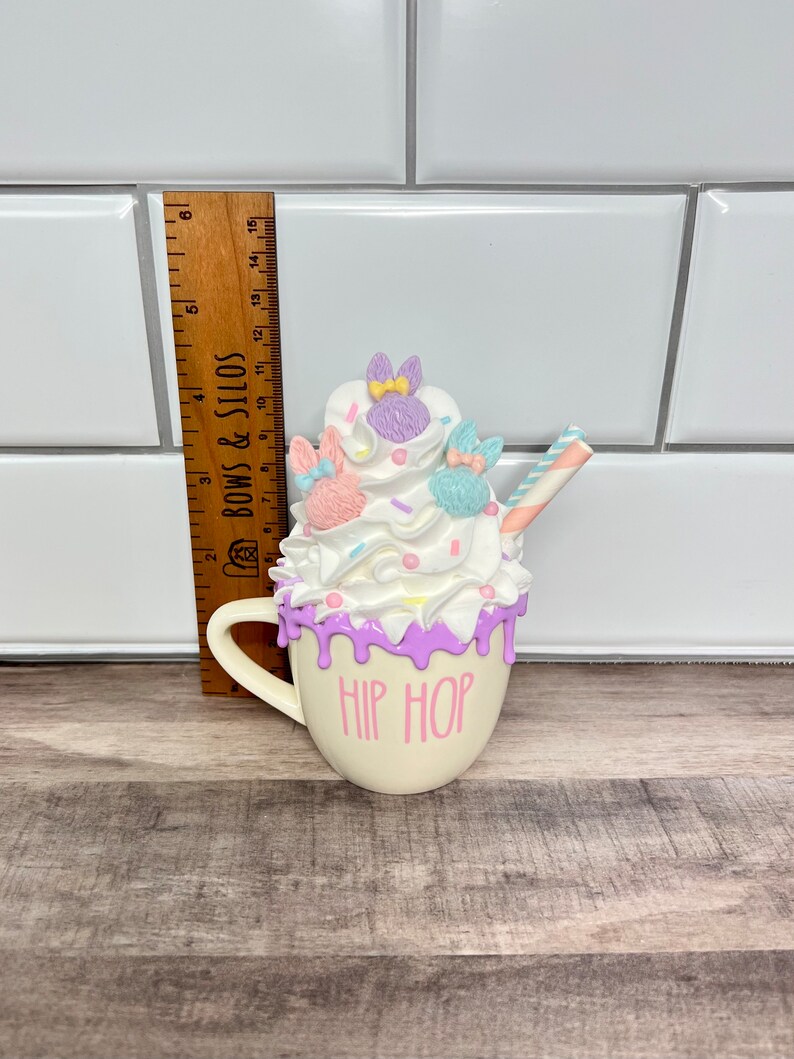 K&ouml;nnte beinhalten: Eine wei&szlig;e Keramiktasse mit rosa und lila Frosting-Design und einem gestreiften Strohhalm. Auf der Tasse steht "HIP HOP" geschrieben.