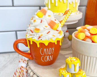 May include: Orange and yellow candy corn themed mug with white whipped cream and candy corn sprinkles. The mug says "Candy Corn" in black lettering. The mug is on a tiered tray with other candy corn themed decorations.