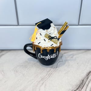 May include: Black coffee mug with gold dripping glaze and the word "Congrats!" written on the side. The mug is topped with a white whipped cream frosting with a black graduation cap, a gold tassel, and a gold diploma.
