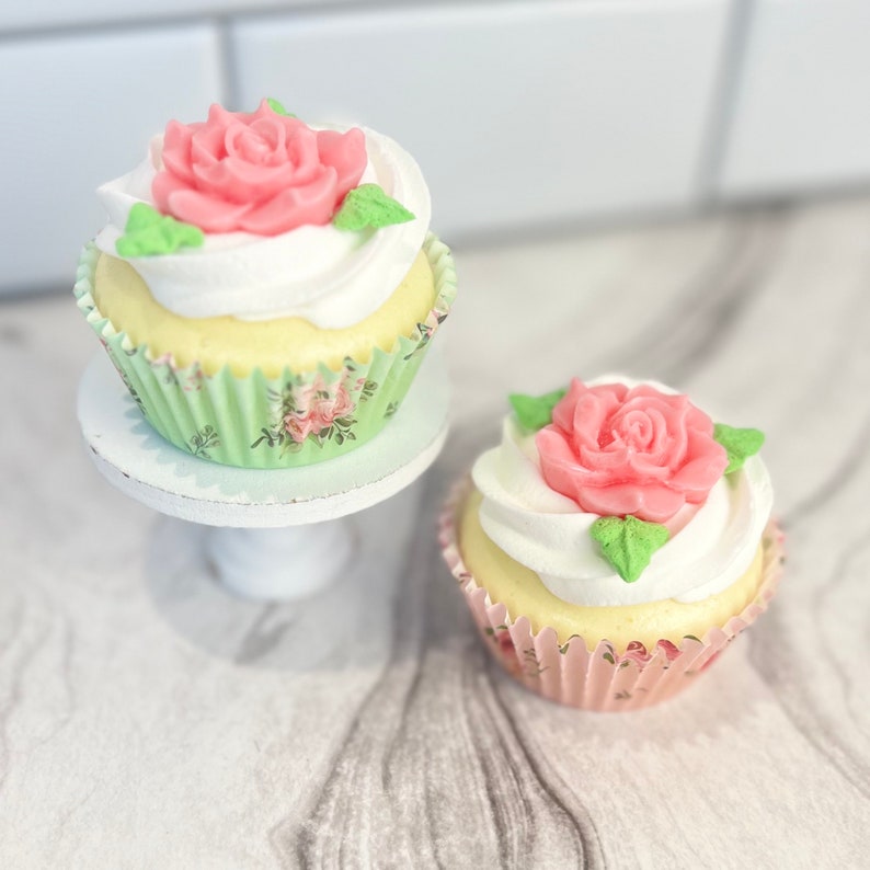 May include: Two cupcakes decorated with pink roses and white frosting. The cupcakes are in green and pink cupcake liners. One cupcake is on a white cake stand.