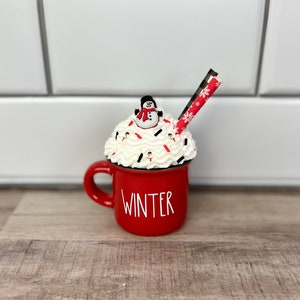 Op de afbeelding: Rode keramische mok met witte letters die "WINTER" zeggen. De mok is gevuld met witte slagroom, rode en witte snoepstokken en een sneeuwpopfiguur.