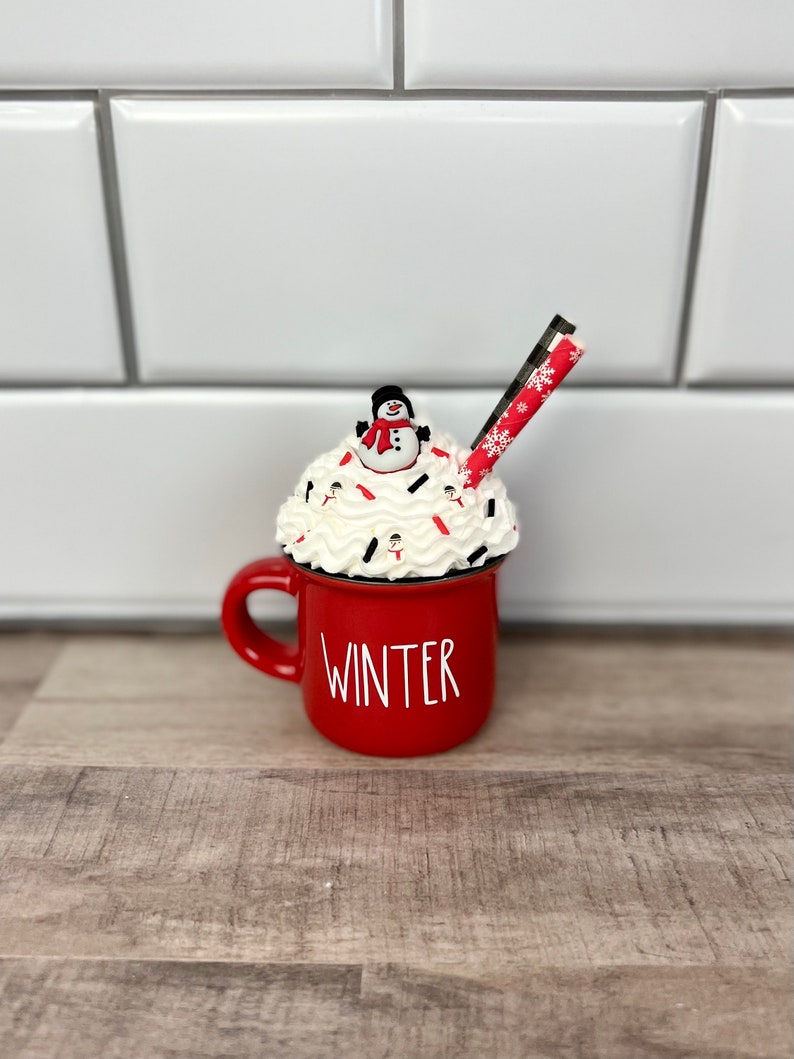 Op de afbeelding: Rode keramische mok met witte letters die "WINTER" zeggen. De mok is gevuld met witte slagroom, rode en witte snoepstokken en een sneeuwpopfiguur.