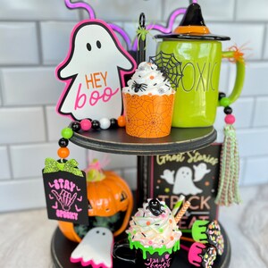 May include: A two-tiered black tiered tray decorated for Halloween. The top tier features a green mug with a witch's hat, a cupcake with a spider web and a spider, a ghost cutout that says "Hey Boo", and a beaded garland. The bottom tier features a black mug with green frosting and a ghost, a pumpkin, a sign that says "Stay Spooky", and a book that says "Ghost Stories Trick or Treat".